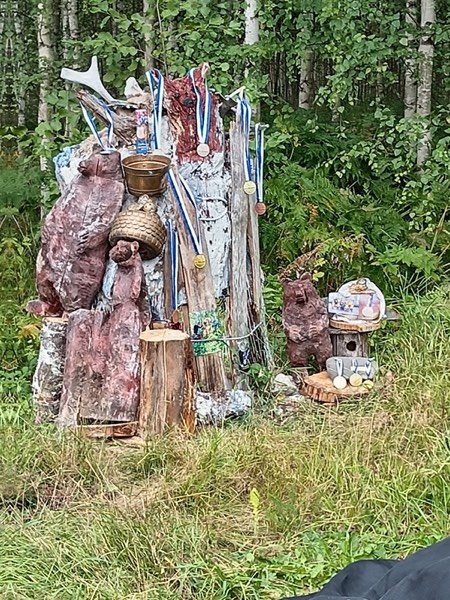 Metsän keskellä oleva taideinstallaatio: kannoista ja oksista koottu rakennelma, johon on ripustettu mitaleja ja pieniä esineitä